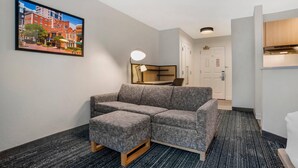 A modern hotel room with a gray sectional sofa, a small ottoman, a framed cityscape picture, a desk with a lamp, and a kitchenette. - MainStay Suites (Tallahassee)