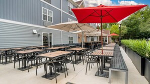 An outdoor patio with multiple tables and chairs, red umbrellas, and a pathway lined with greenery. - MainStay Suites (Tallahassee)