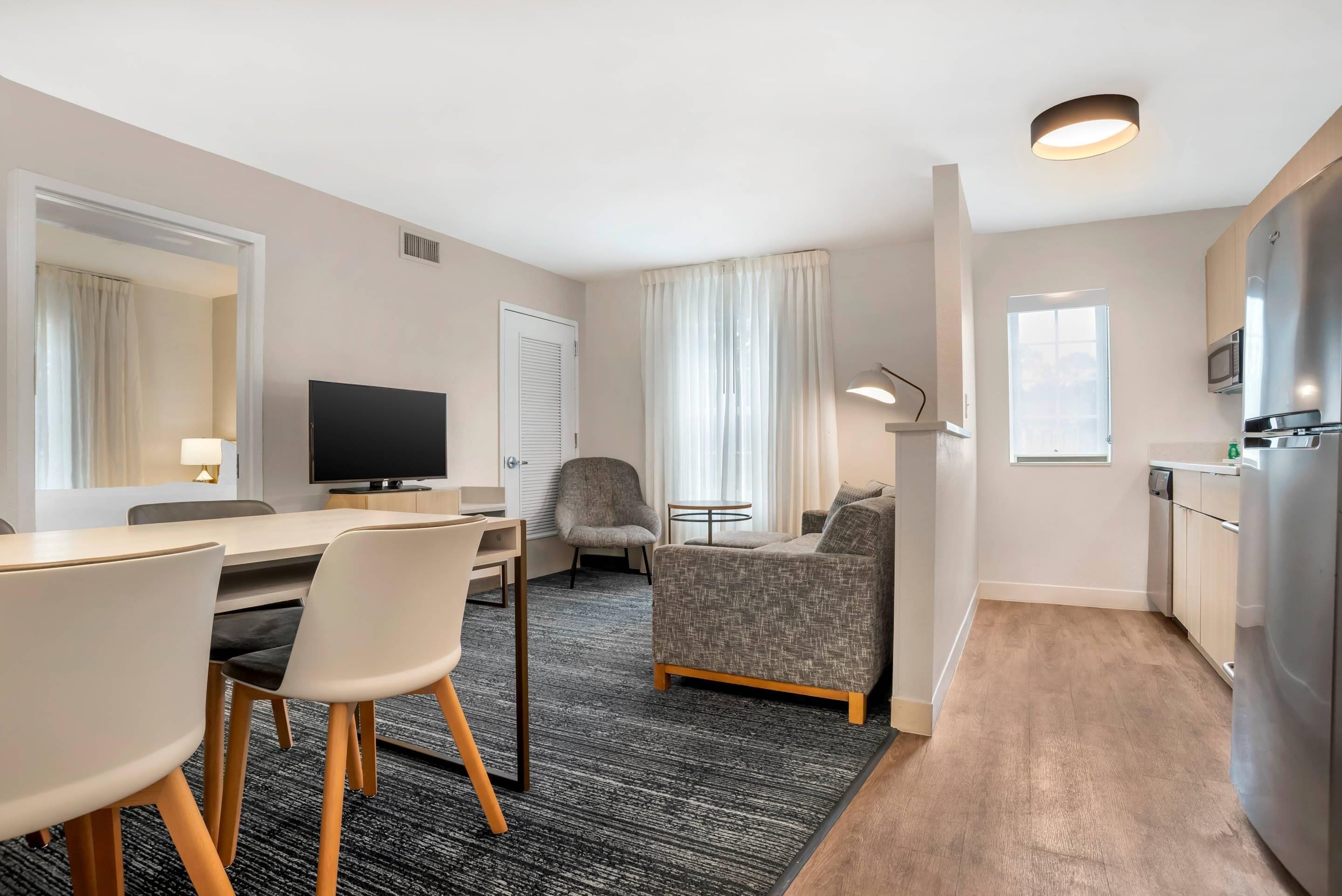 Habitación de hotel con comedor, televisor, sofá, una mesita y una zona de cocina con microondas.