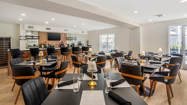 A modern dining area with multiple tables and chairs, a kitchen in the background, and large windows allowing natural light. - MainStay Suites (Tallahassee)