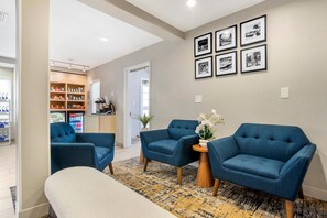 Ein modernes Wohnzimmer mit blauen Sesseln, einem Couchtisch und einer Bank. An der Wand befinden sich gerahmte Bilder und ein mit Getränken gefüllter Kühlschrank.