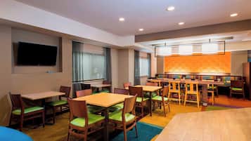 A hotel room with a dining area, a flat-screen TV mounted on the wall, and a large window with curtains.