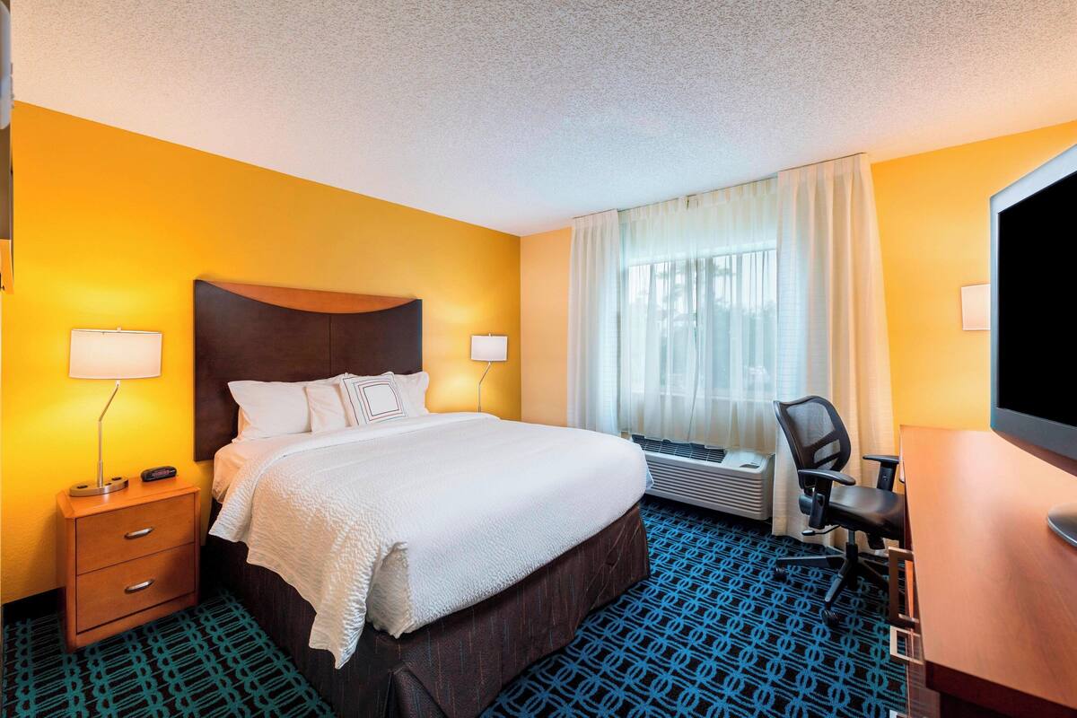 A hotel room with a large bed, bedside lamps, a desk with a chair, and a large window with curtains.