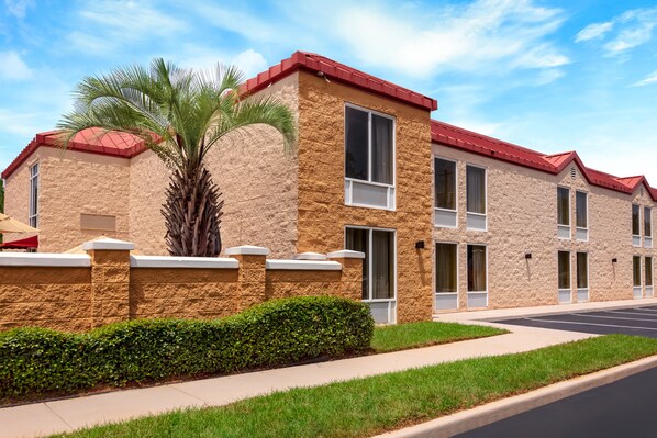 A multi-story building with a stone facade and red roof, featuring a parking lot and a sidewalk with a palm tree.