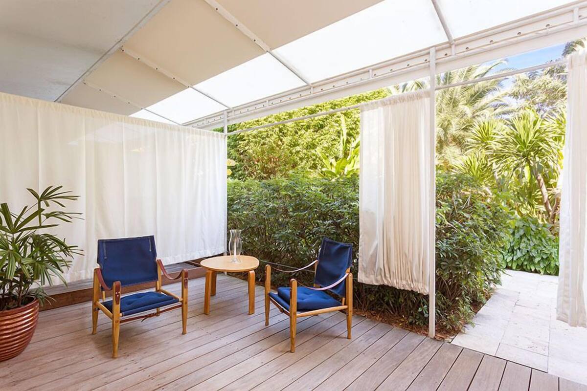 Un espace extérieur aménagé avec des sièges, abrité par un toit, comprenant deux chaises bleues et une petite table, le tout entouré de rideaux blancs et d’une végétation luxuriante.