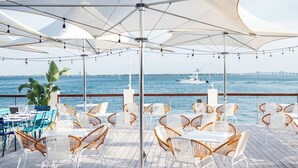 Un área de comedor al aire libre con sillas y mesas de mimbre blanco, grandes sombrillas blancas y vista al océano y un puente en el fondo.