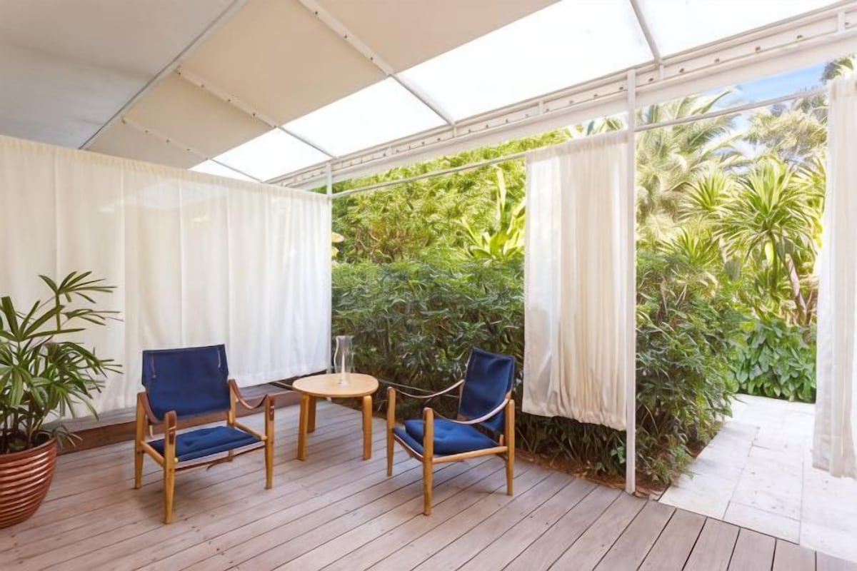 A covered outdoor seating area with wooden chairs and a table, surrounded by white curtains and lush greenery.