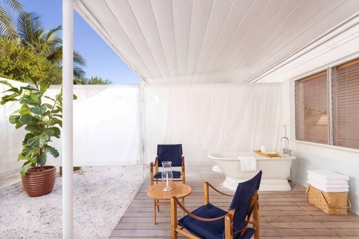 Un espace extérieur abrité, doté d’une terrasse en bois, de deux chaises, d’une petite table, d’une baignoire et d’une plante en pot.