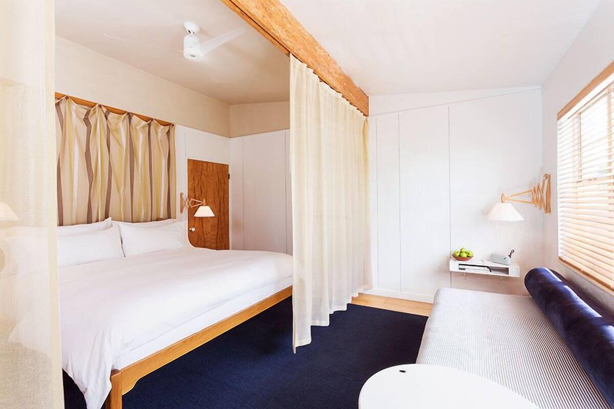 Une chambre d’hôtel comprenant un lit, une table de chevet avec une lampe, un petit coin salon avec un coussin et une fenêtre avec des stores.