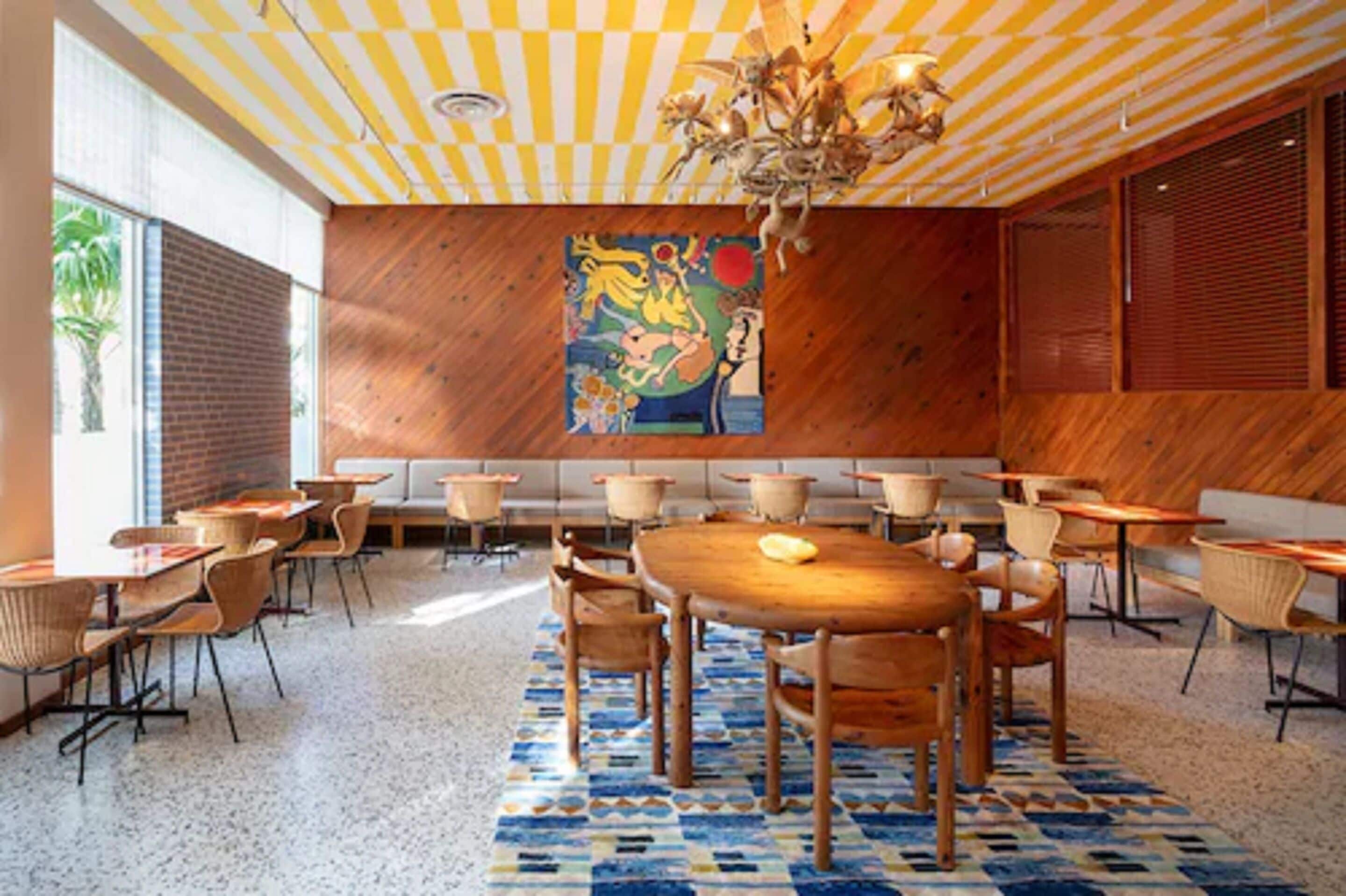 A dining area with wooden tables and chairs, a large mural on the wall, and a patterned rug.