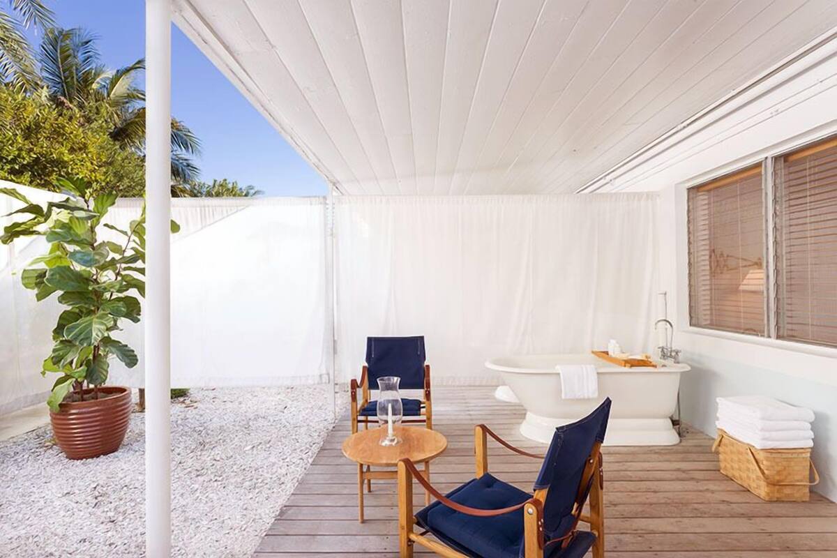 Un espace extérieur abrité doté d’une terrasse en bois, de deux chaises, d’une table, d’une plante en pot, d’une baignoire blanche et d’une fenêtre avec des stores.