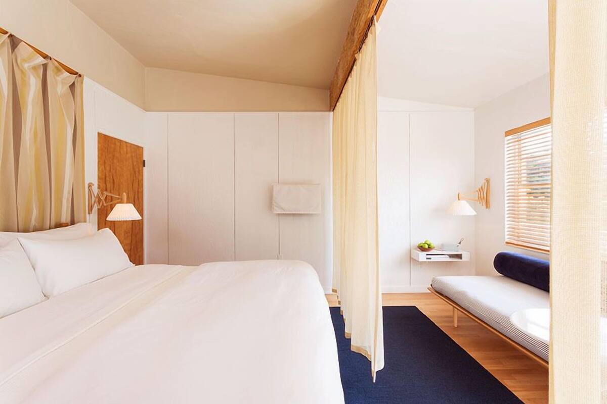 Une chambre d’hôtel avec un grand lit, une table de chevet avec une lampe, un petit coin salon avec un banc et une fenêtre avec des stores.