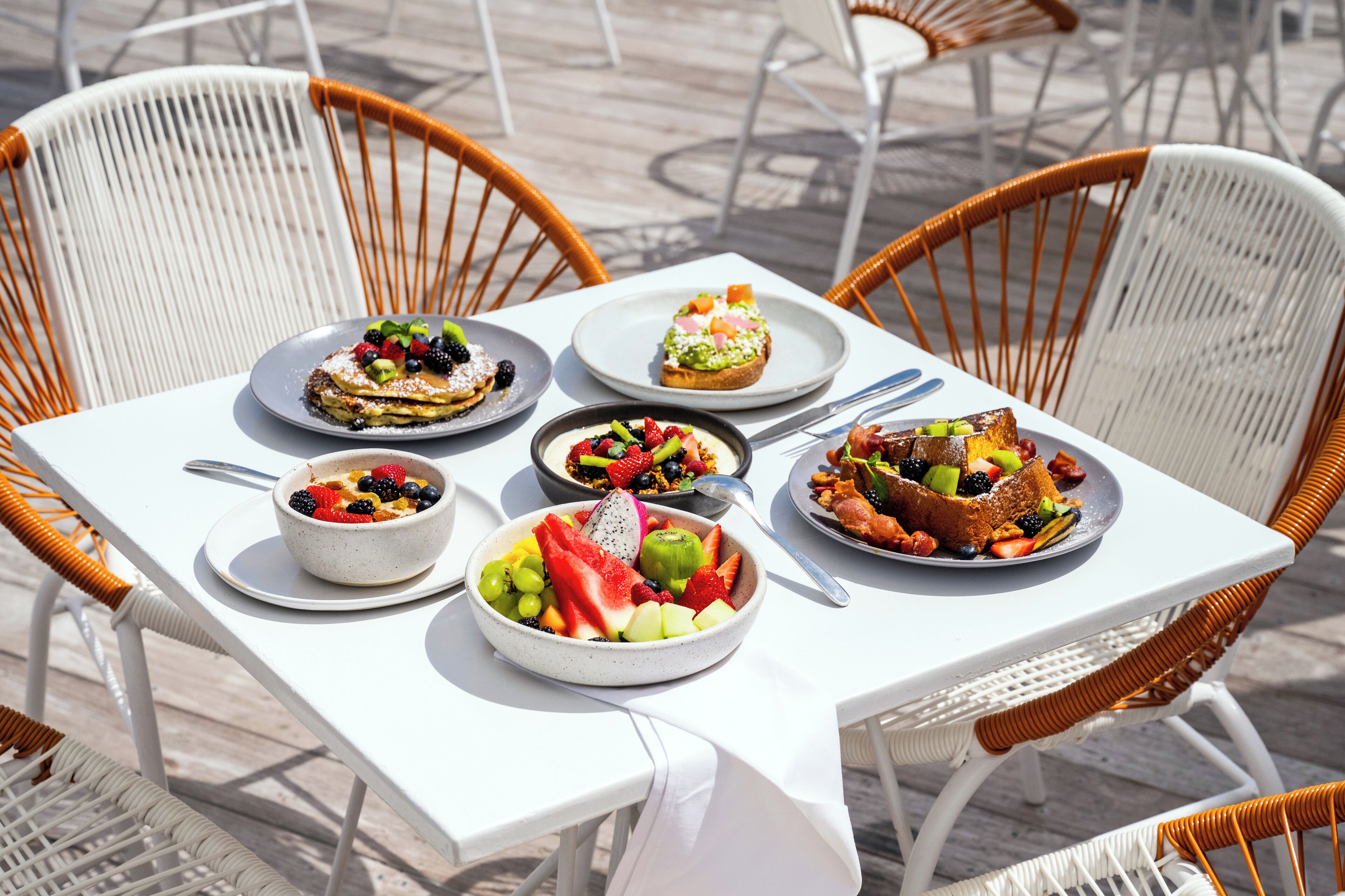 A table with various dishes, including a fruit bowl, a dessert, and a plate with a cake, surrounded by wicker chairs.