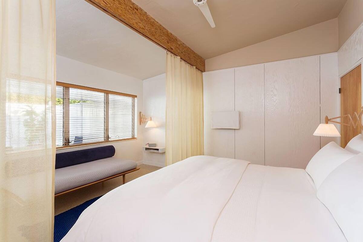 Une chambre d’hôtel avec un grand lit, un banc, une fenêtre avec des stores, une table de chevet avec une lampe et une poutre en bois au plafond.