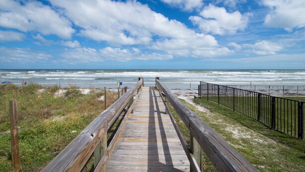 Une promenade en bois menant à une plage, avec un ciel dégagé et une vue sur l’océan.
