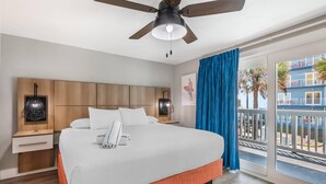 Une chambre d’hôtel comprenant un lit, un ventilateur de plafond, une table de chevet, un téléphone, une fenêtre donnant sur un immeuble et un balcon avec des rideaux bleus.