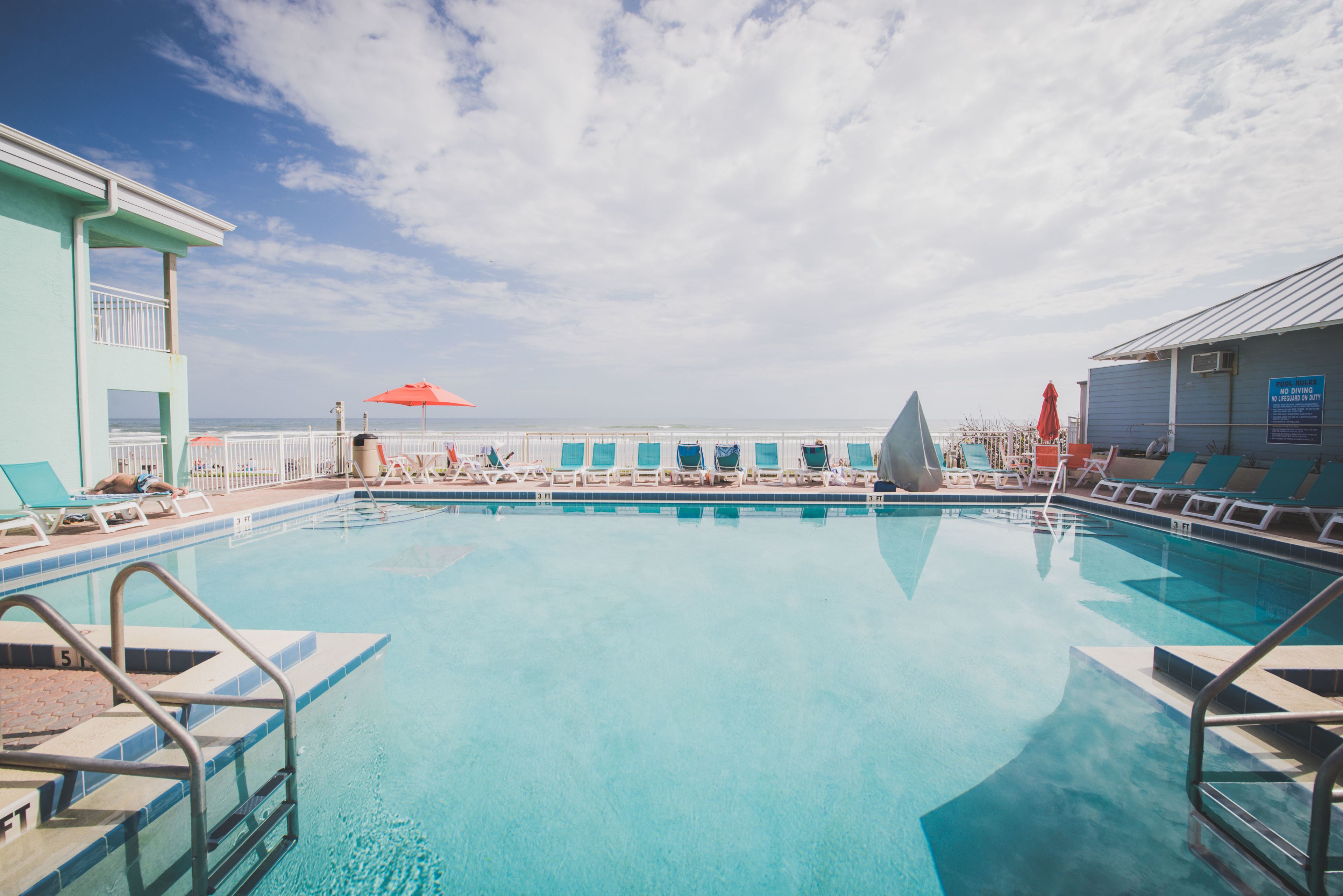 A swimming pool with clear blue water, surrounded by lounge chairs and umbrellas, with a view of the ocean and a building in the background.
