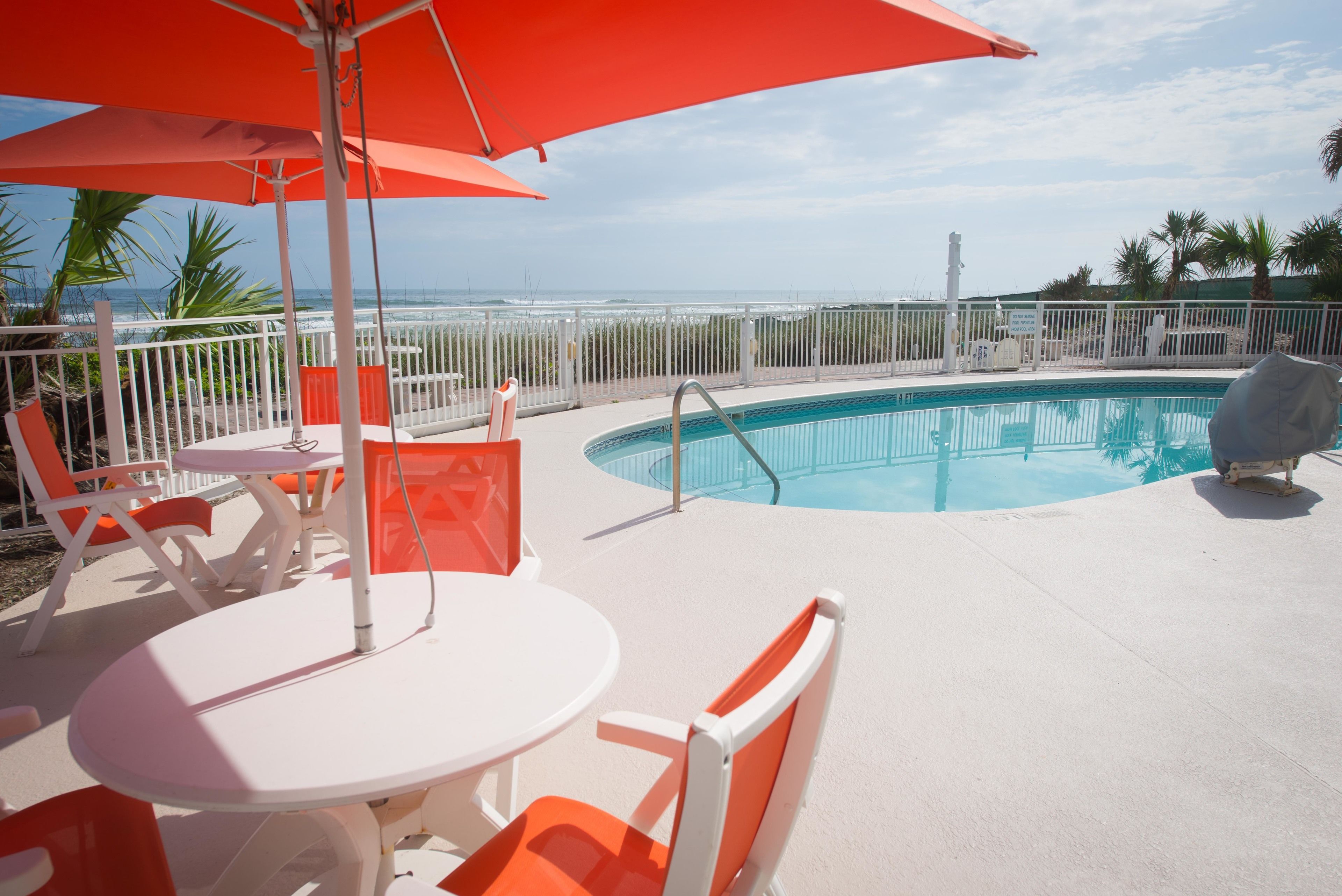 A poolside area with a pool, white and orange chairs, and a red umbrella.