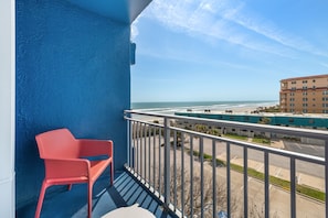 A balcony with a red chair, a view of the beach, and a parking lot.