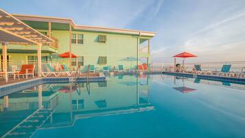 A swimming pool with lounge chairs and umbrellas, a building with a balcony, and a clear sky.
