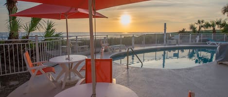 A poolside area with a table and chairs, a red umbrella, and a view of the sunset over the water.