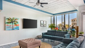 Un salon doté d’une grande fenêtre, d’un téléviseur à écran plat, d’un canapé, d’un pouf et d’un ventilateur de plafond.