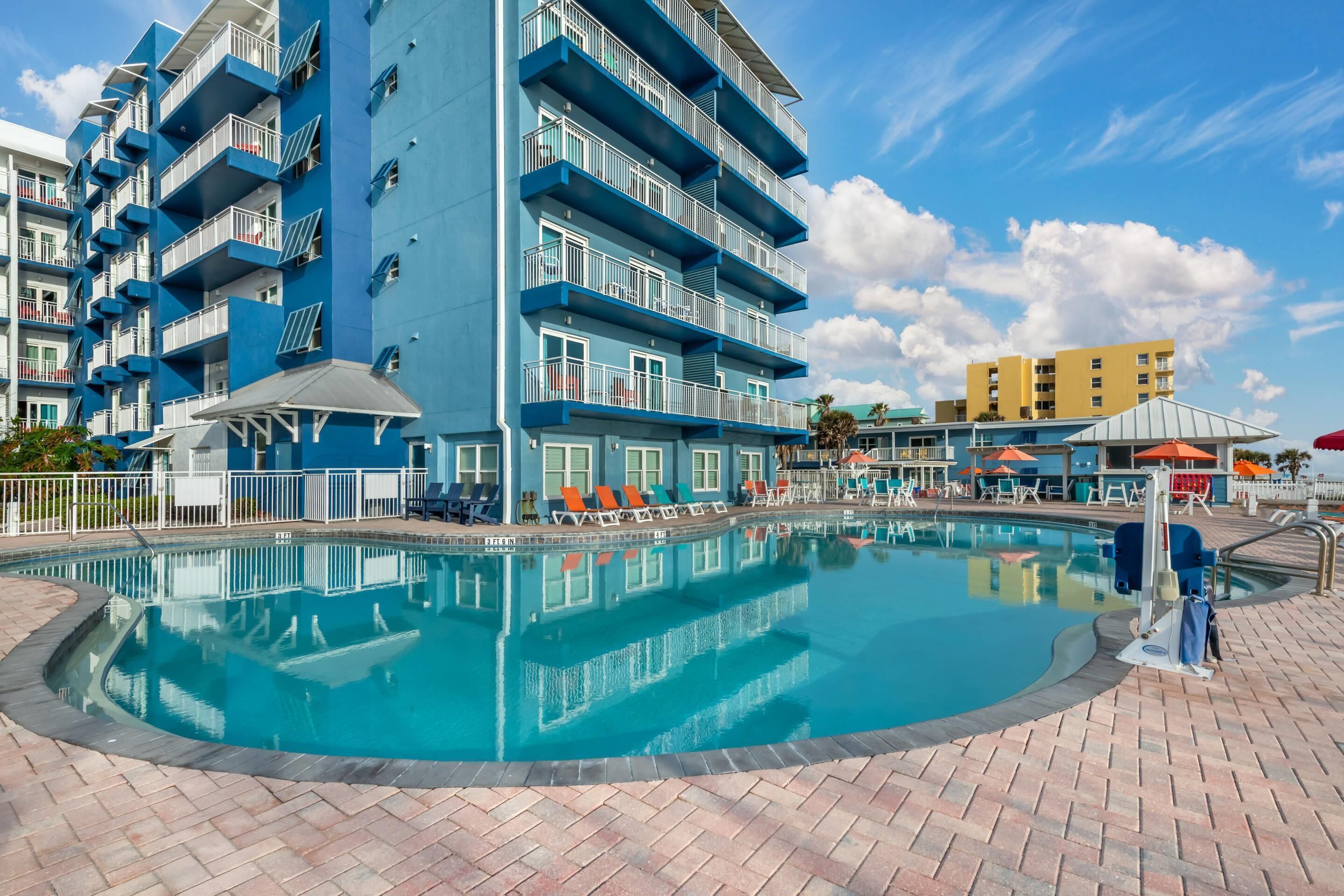 A swimming pool surrounded by a multi-story building with balconies, a brick walkway, and a clear sky.