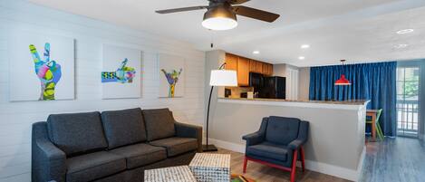 A modern living room with a gray sofa, a blue armchair, and a colorful rug. There is a kitchen area with a microwave and a bar counter.