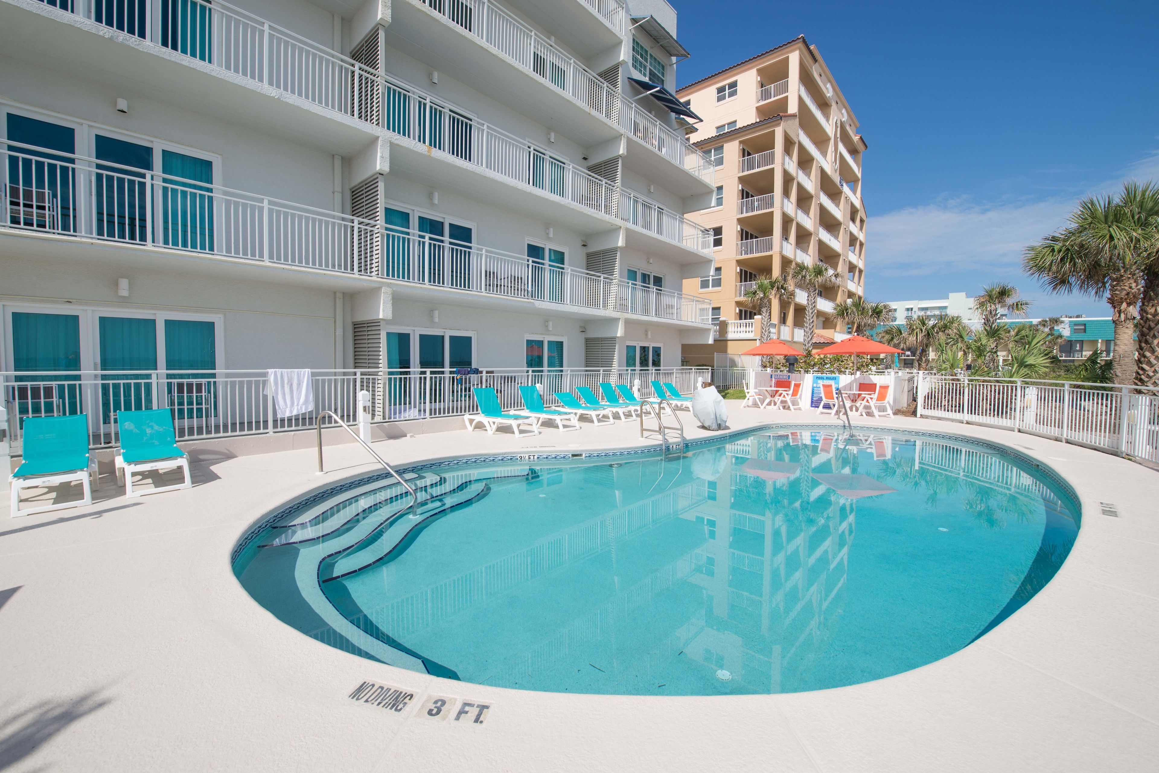 A swimming pool with lounge chairs and umbrellas, surrounded by a white building with balconies.