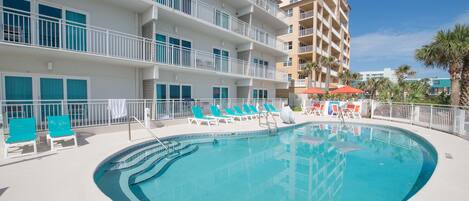 A swimming pool with lounge chairs and umbrellas, surrounded by a white building with balconies.