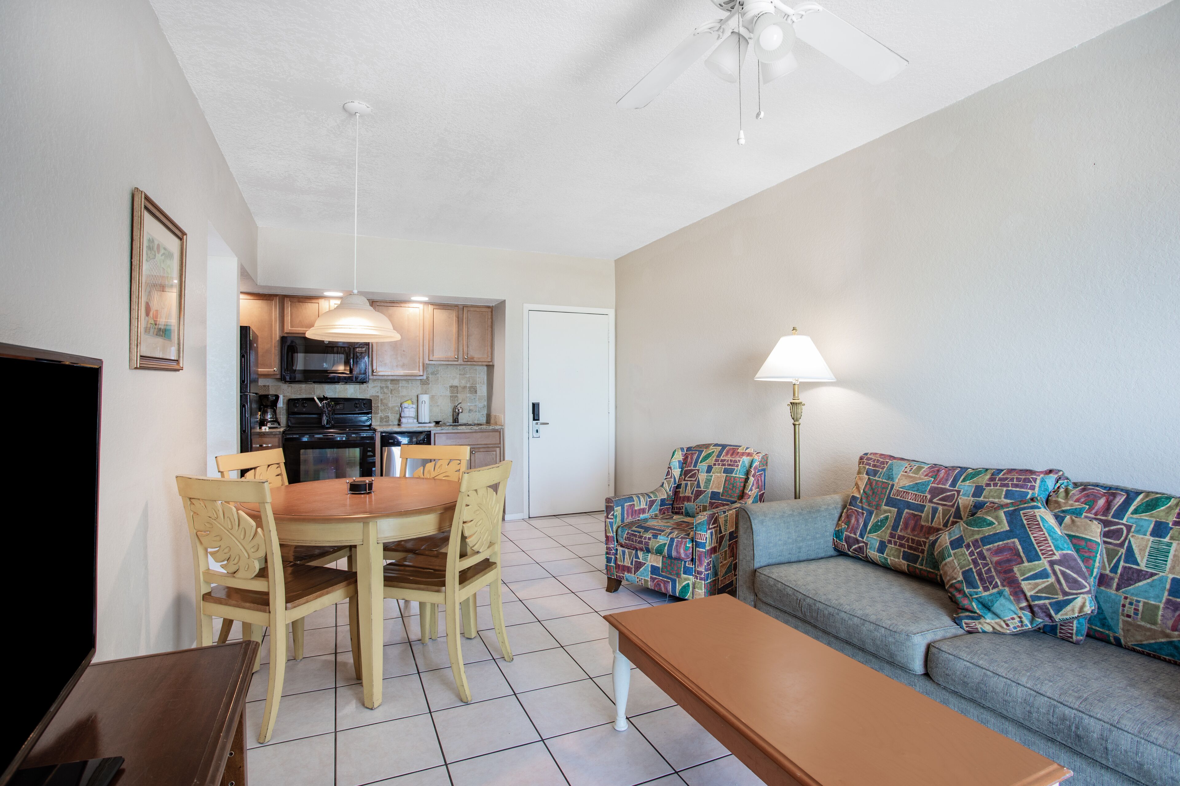 A living room with a sofa, a coffee table, and a dining table with chairs. There is a kitchen in the background with a microwave and a refrigerator.
