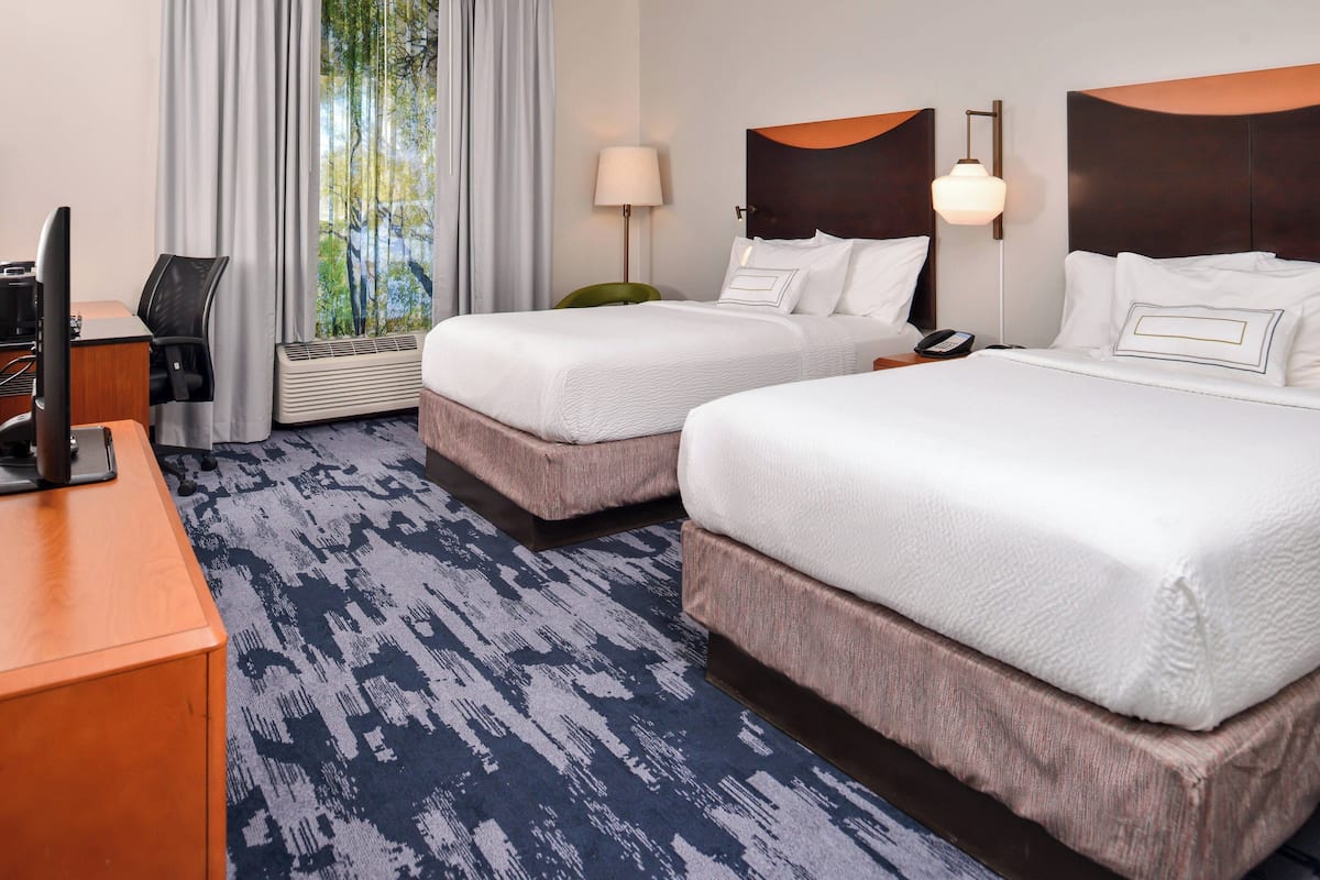 A hotel room with two beds, a desk, a chair, a lamp, and a window with curtains.