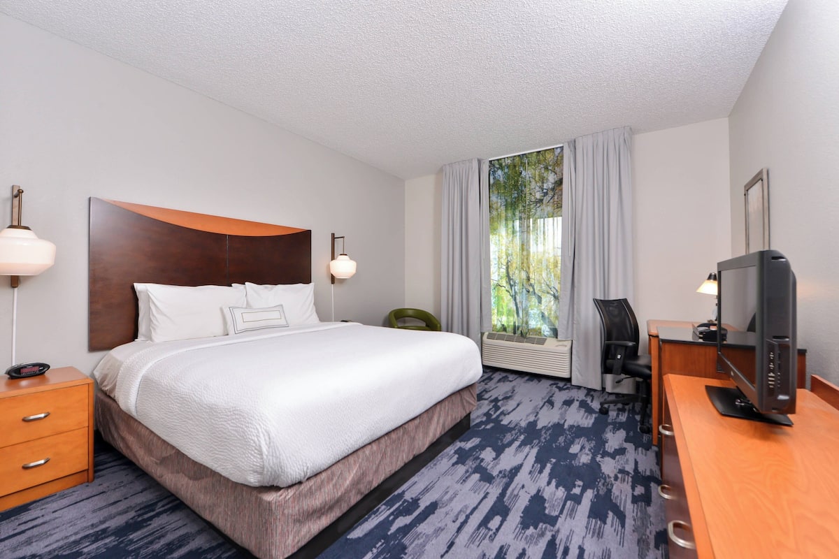 A hotel room with a large bed, a desk with a television, a chair, a desk lamp, and a window with curtains.