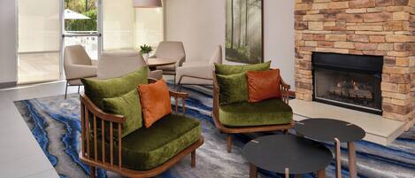 A modern living room with a stone fireplace, two green armchairs with orange cushions, a round coffee table, and a large window with a view of a deck and greenery.