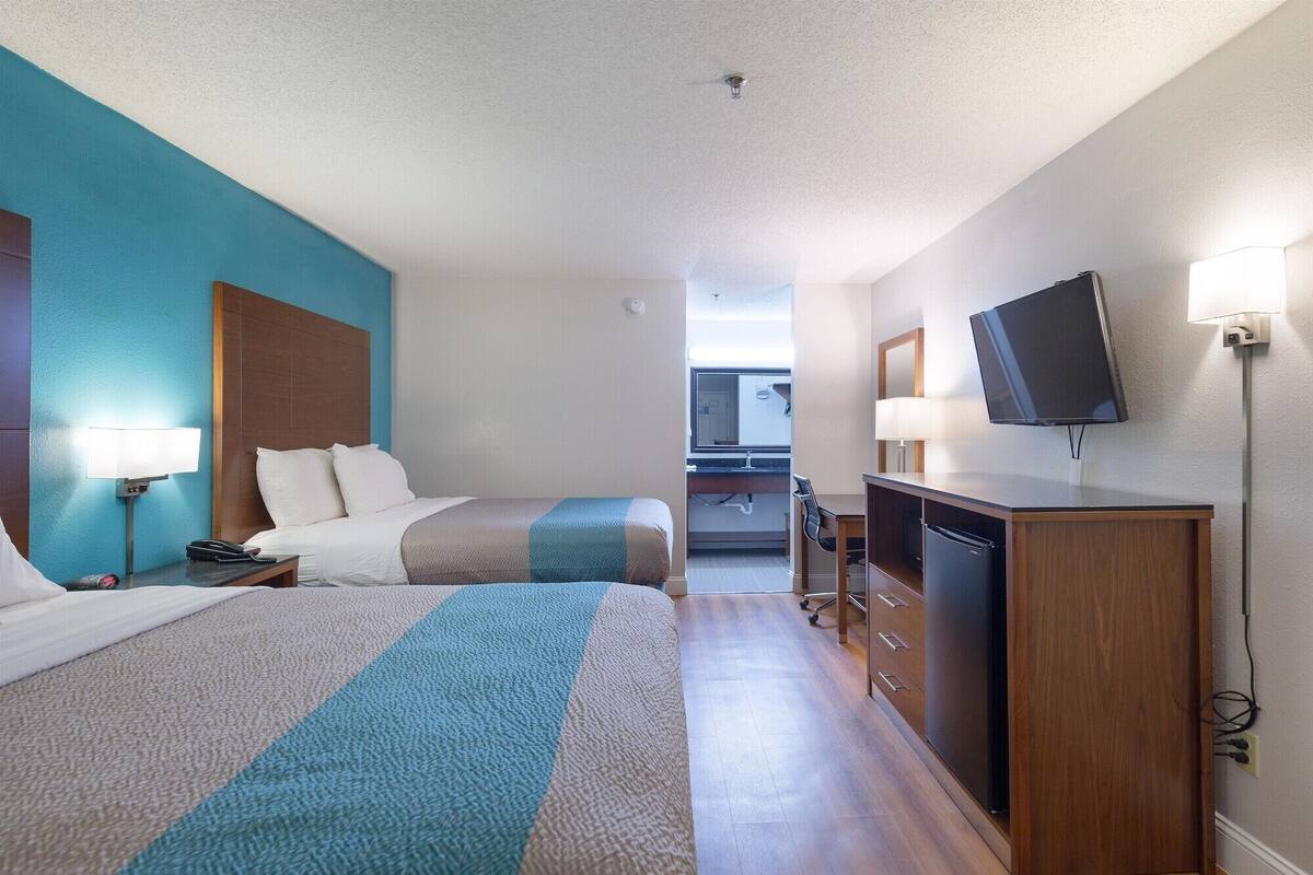 A hotel room with two beds, a desk, a TV, and a bathroom visible in the background.