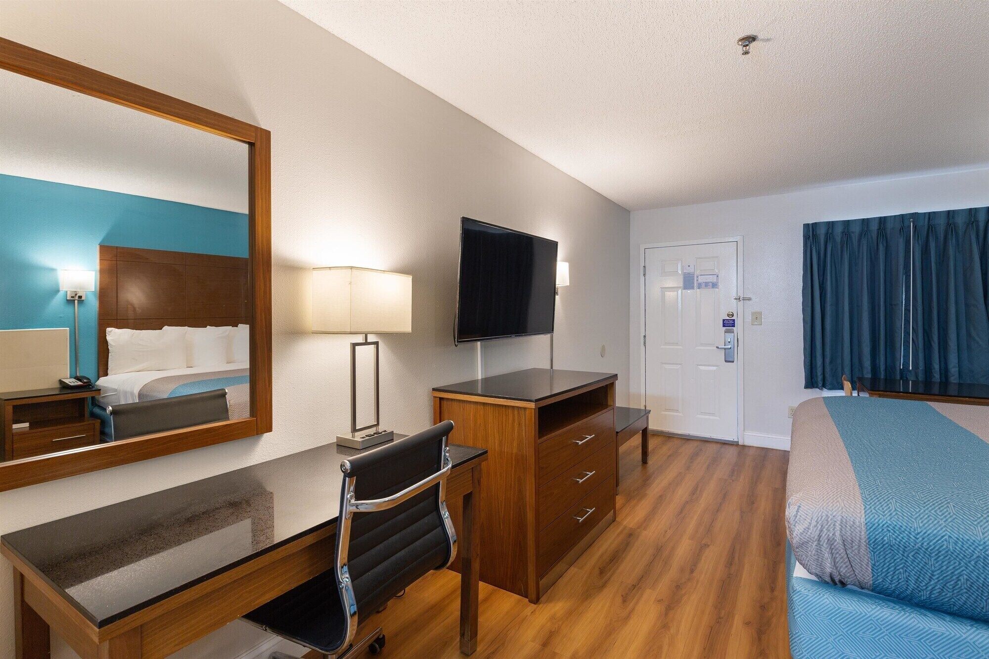 A hotel room with a bed, a desk with a chair, a television, a mirror, and a door.