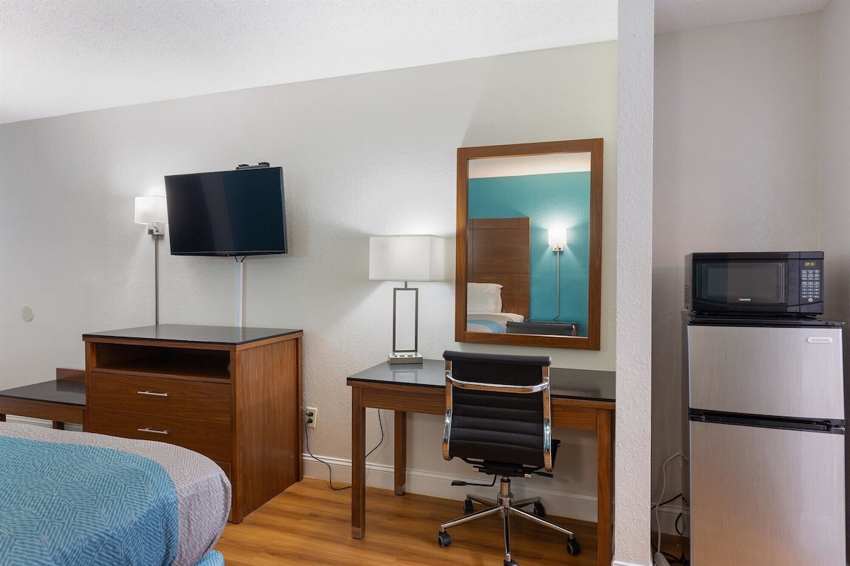 A hotel room with a bed, a desk, a chair, a television, a mirror, a microwave, and a refrigerator.
