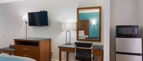 Una habitación de hotel con cama, escritorio, silla, televisión, espejo, microondas y refrigerador.