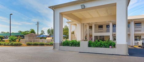 A two-story building with a covered outdoor area, a parking lot, and a green-roofed building in the background.