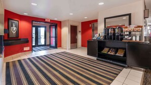 A hotel lobby with a red wall, an exit sign, a coffee station, and a striped carpet. - Extended Stay America Suites Chicago Romeoville Bollingbrook (Romeoville)