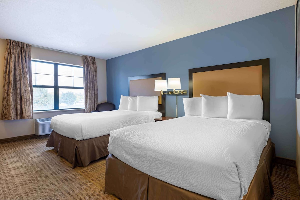 A hotel room with two beds, a large window with curtains, a desk, and a lamp.
