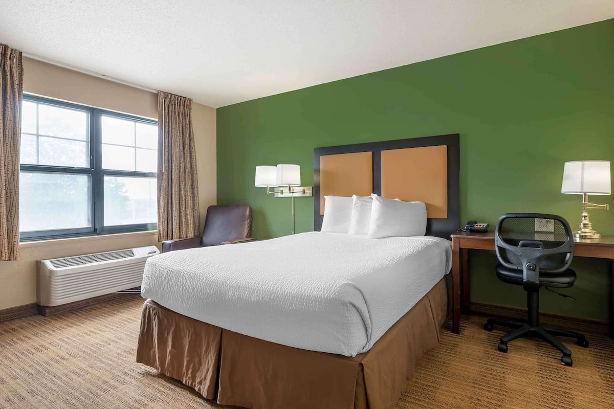 A hotel room with a large bed, a desk with a chair, a window with curtains, and a wall-mounted lamp.