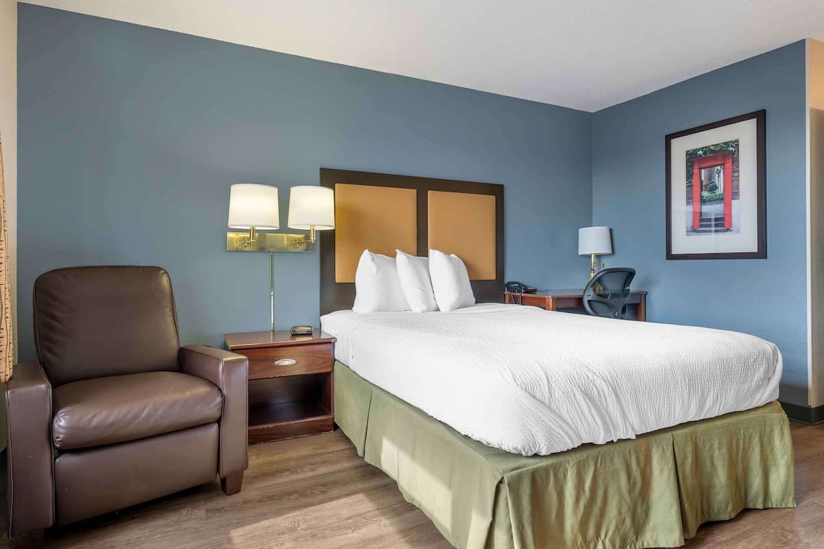 A hotel room with a bed, a chair, a nightstand, a lamp, and a framed picture on the wall.