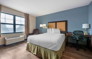 A hotel room with a large bed, a desk with a chair, a window with curtains, and a wall-mounted lamp. - Extended Stay America Suites Chicago Romeoville Bollingbrook (Romeoville)