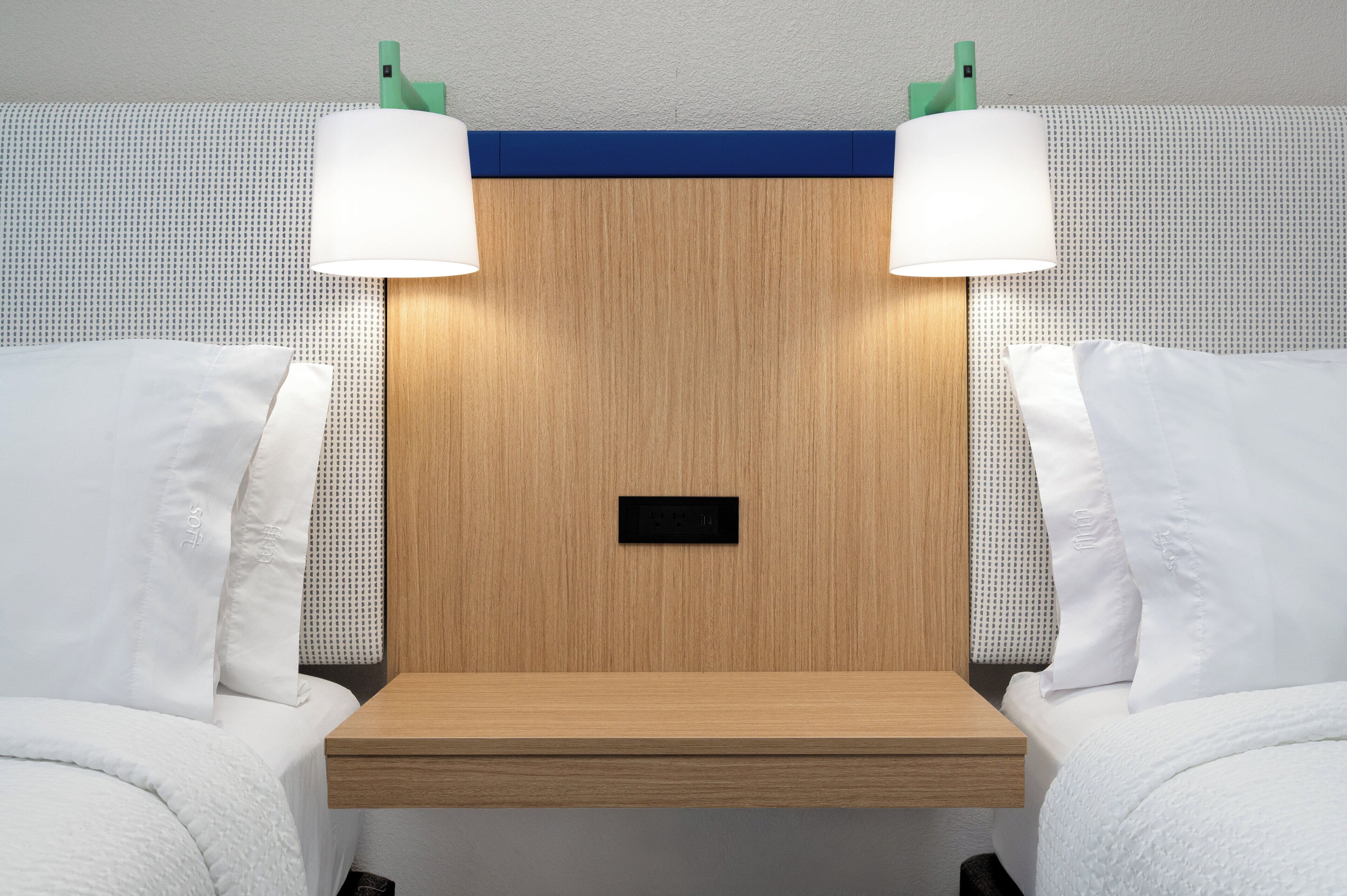 A hotel room with a wooden headboard, two white pillows, a small wooden nightstand, and two wall-mounted lamps.