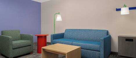 Una habitación con un sofá azul, un sillón verde, una mesa de centro de madera, un cubo de basura rojo y una lámpara de pie verde.