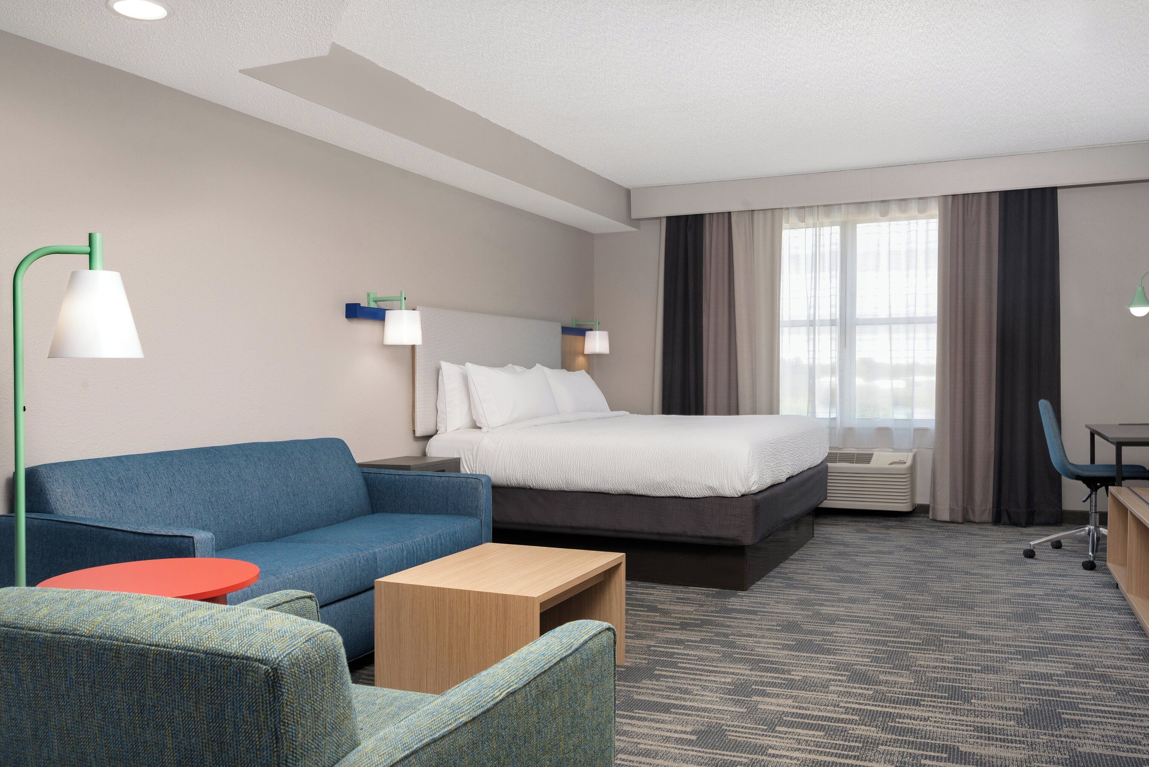 A hotel room with a bed, a blue sofa, a red table, a wooden side table, a desk with a chair, and a window with curtains.