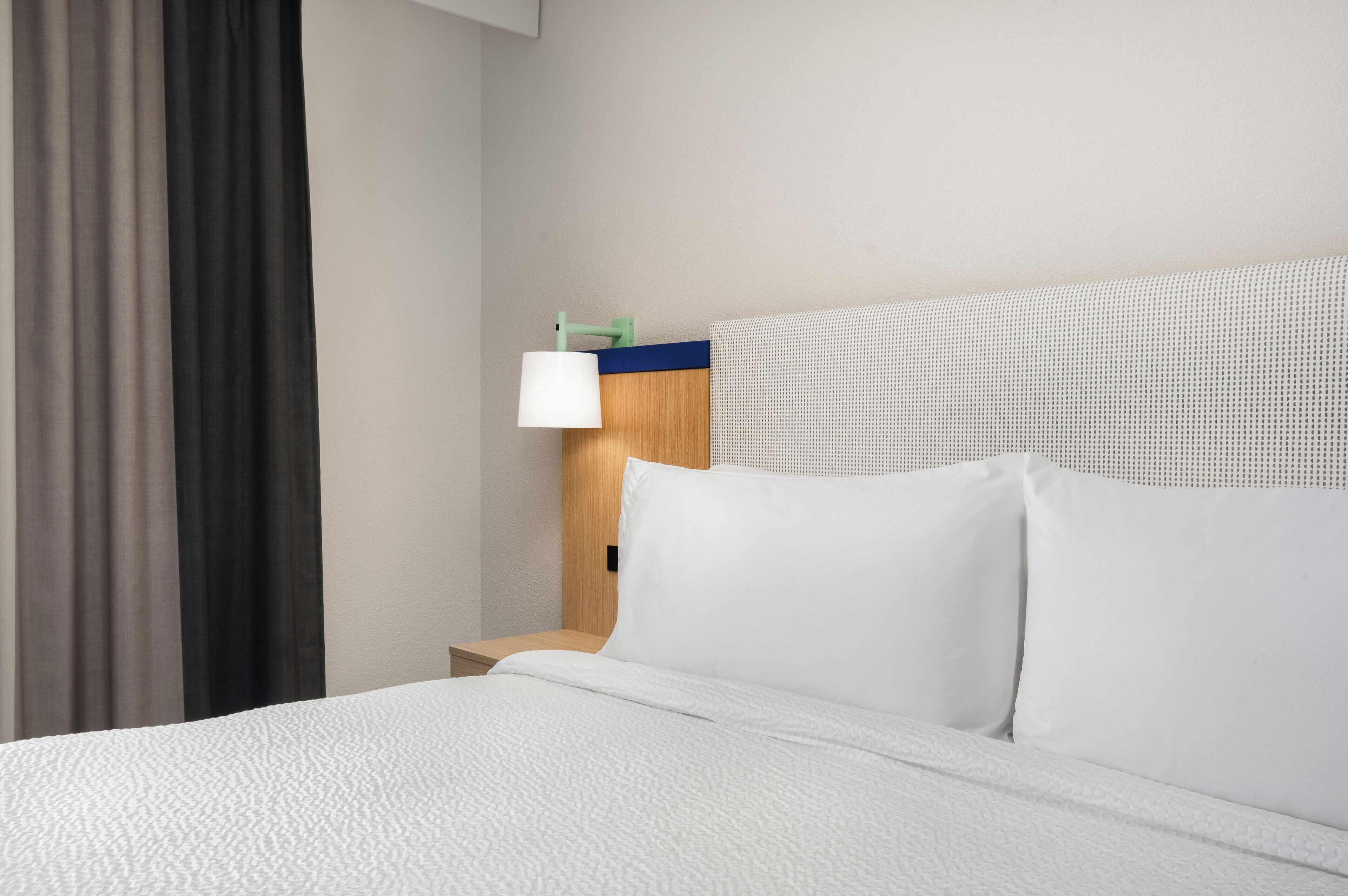 Una cama bien hecha con sábanas blancas, un cabecero de madera y una mesita de noche con una lámpara.