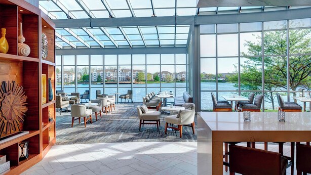 Amplio espacio interior con una gran pared de vidrio que ofrece vistas a un cuerpo de agua y edificios. Varias zonas de estar con mesas y sillas.
