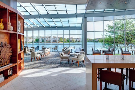 A spacious indoor area with a large glass wall offering a view of a body of water and buildings. There are multiple seating areas with tables and chairs.